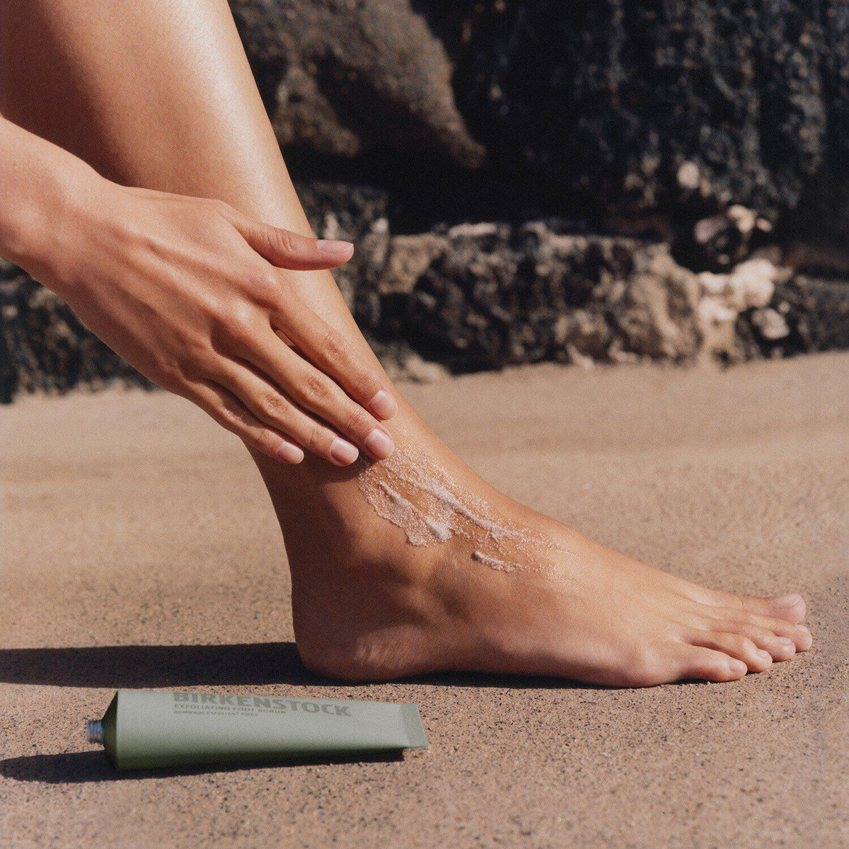 Person applying Birkenstock exfoliating foot scrub to ankle outdoors on sandy surface