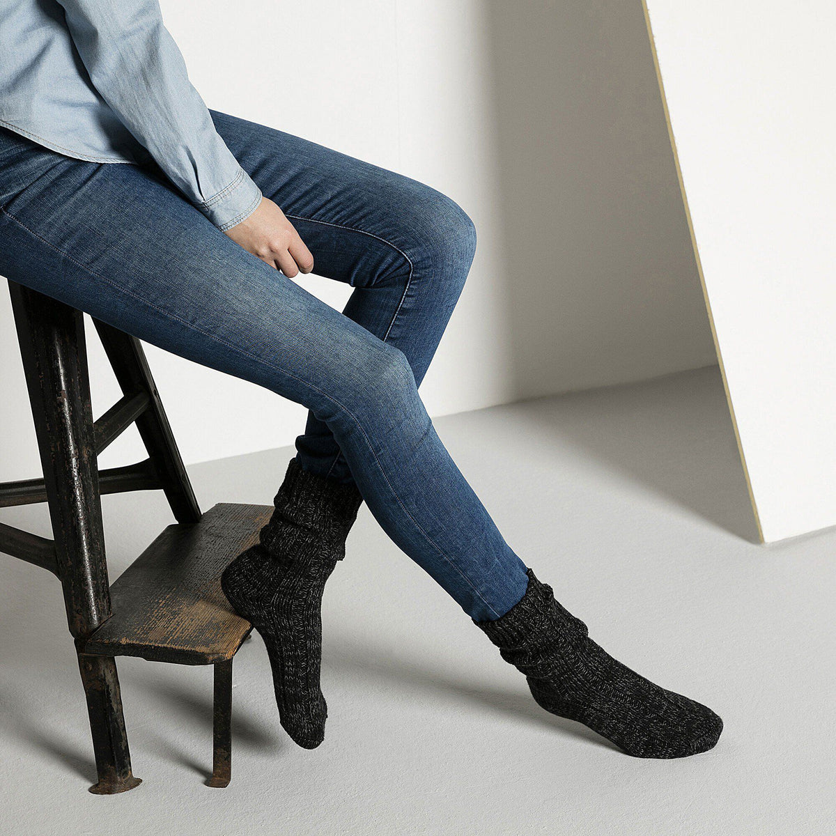 Model seated on rustic stool wearing dark knitted Birkenstock cotton twist socks with jeans