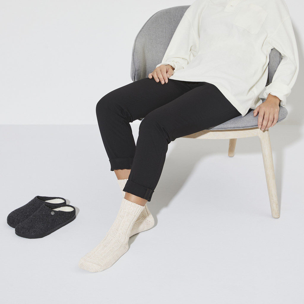 Person seated in chair wearing off-white ribbed ankle socks with dark slippers nearby