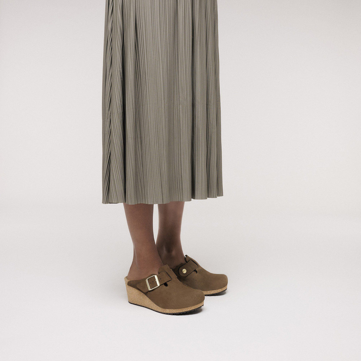 Woman wearing brown suede wedge clogs with gold buckle strap and cork sole
