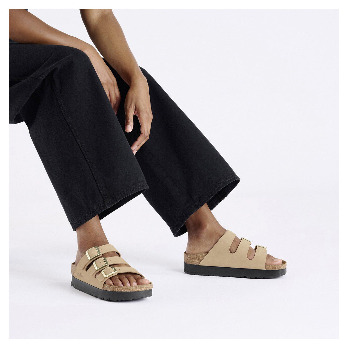 Woman seated wearing three-strap platform sandals with gold buckles and cork footbed