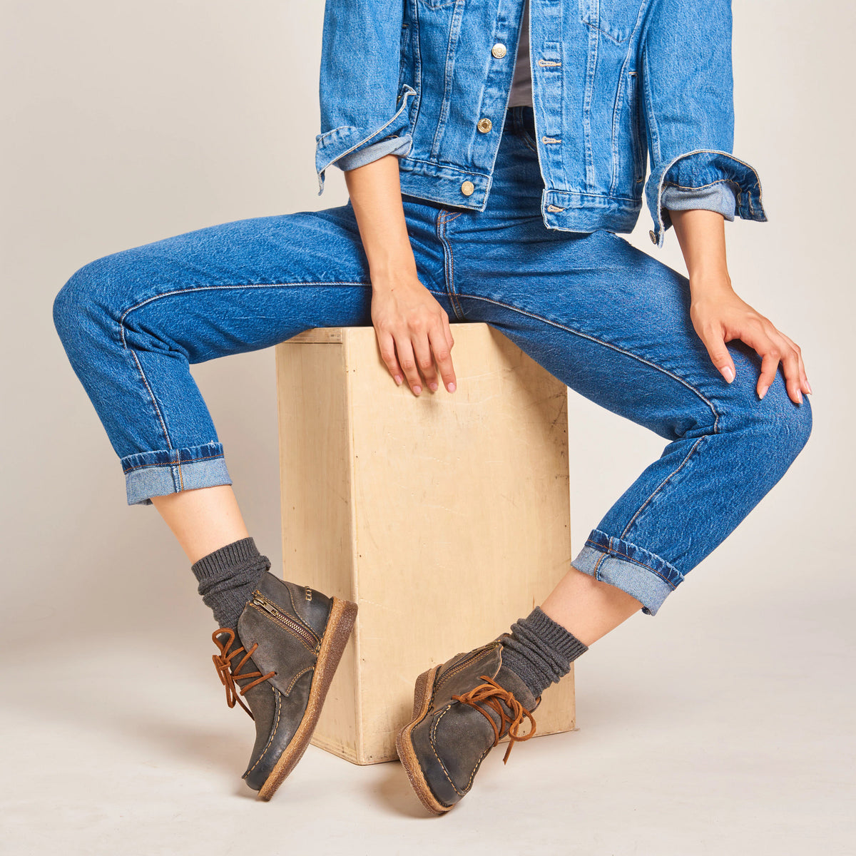 Woman seated on wooden cube wearing lace-up taupe ankle boots with grey knit socks and cuffed jeans