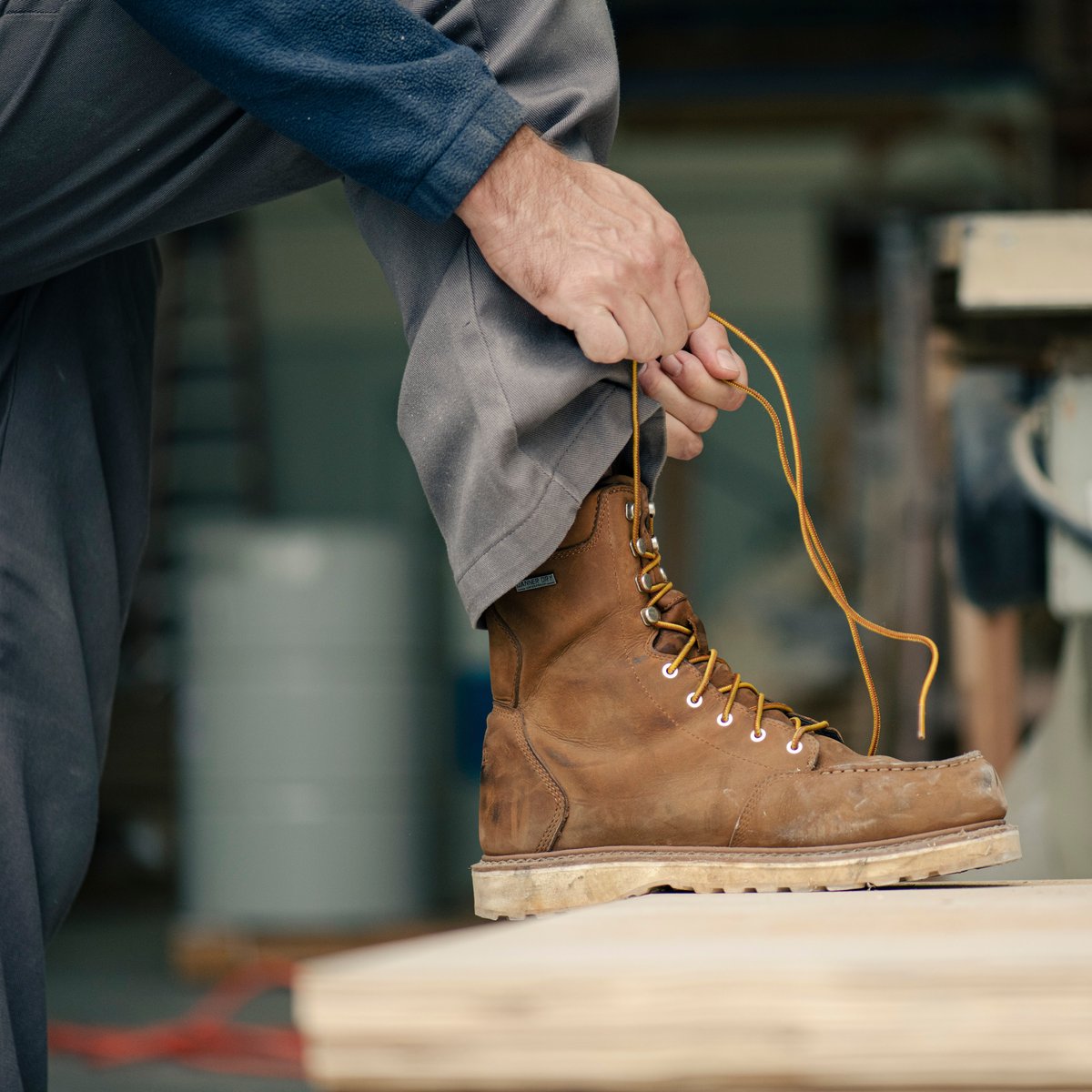 Worker tying laces on a tall brown leather moc toe work boot in an industrial setting