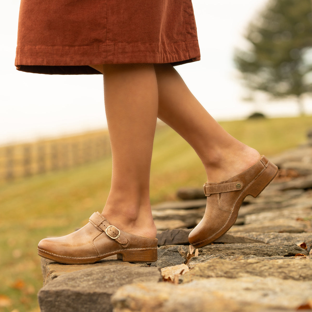 Woman wearing tan leather buckle-strap clogs walking on a stone wall outdoors