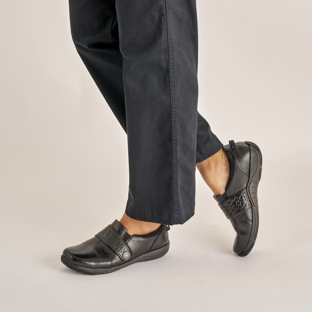 Woman wearing black leather slip-on shoes with decorative strap detail, paired with dark trousers