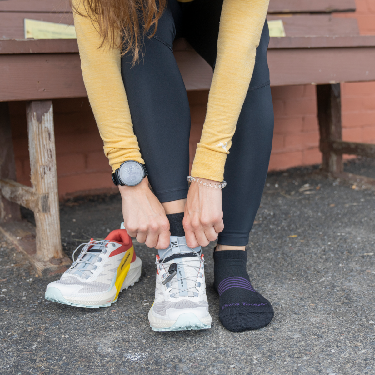 Woman adjusting Darn Tough no-show tab socks while seated on outdoor bench with trail running shoes