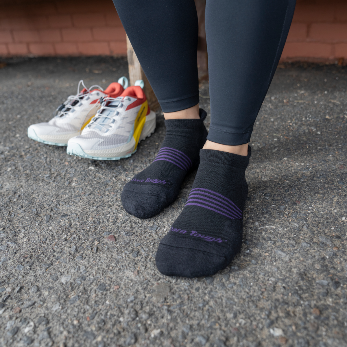 Person wearing black Darn Tough no-show tab socks with purple stripe detail, standing on pavement