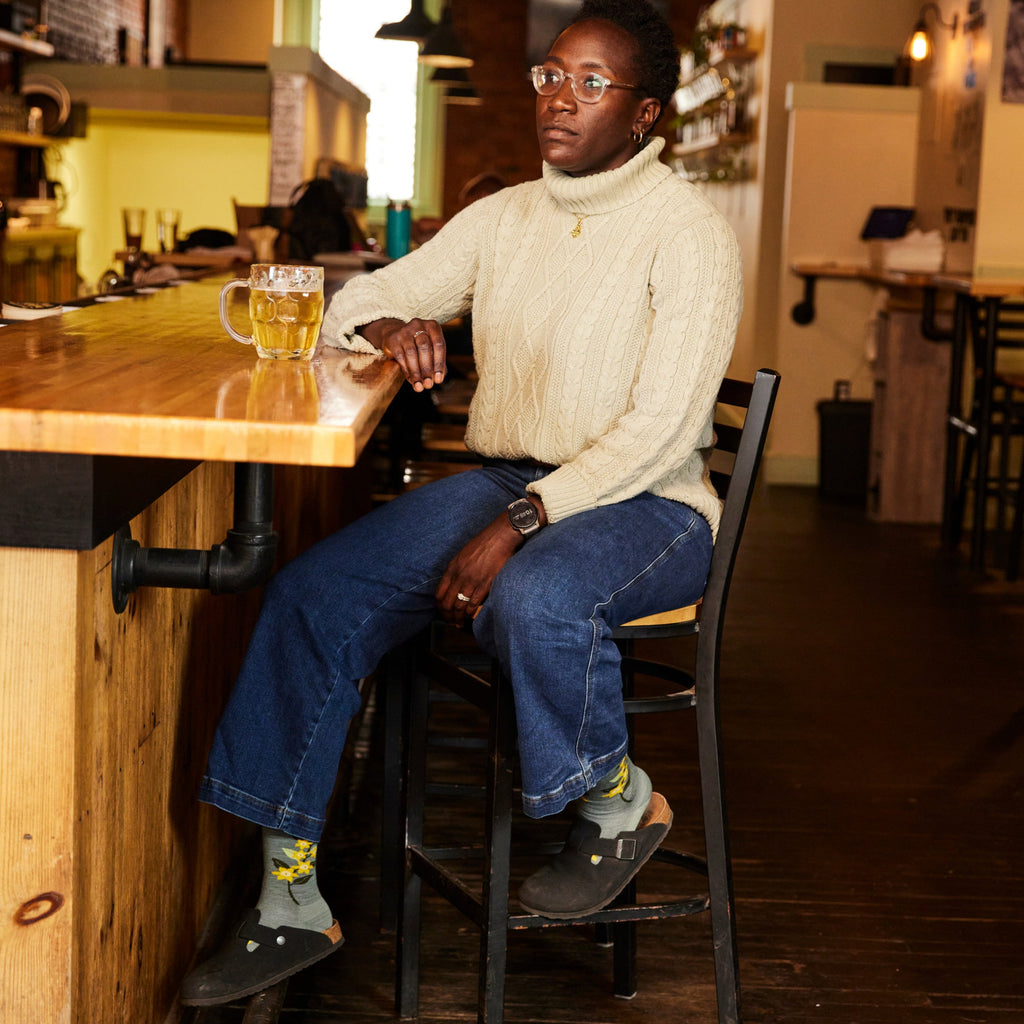 Woman seated at a bar wearing patterned seafoam socks with Birkenstock clogs and jeans