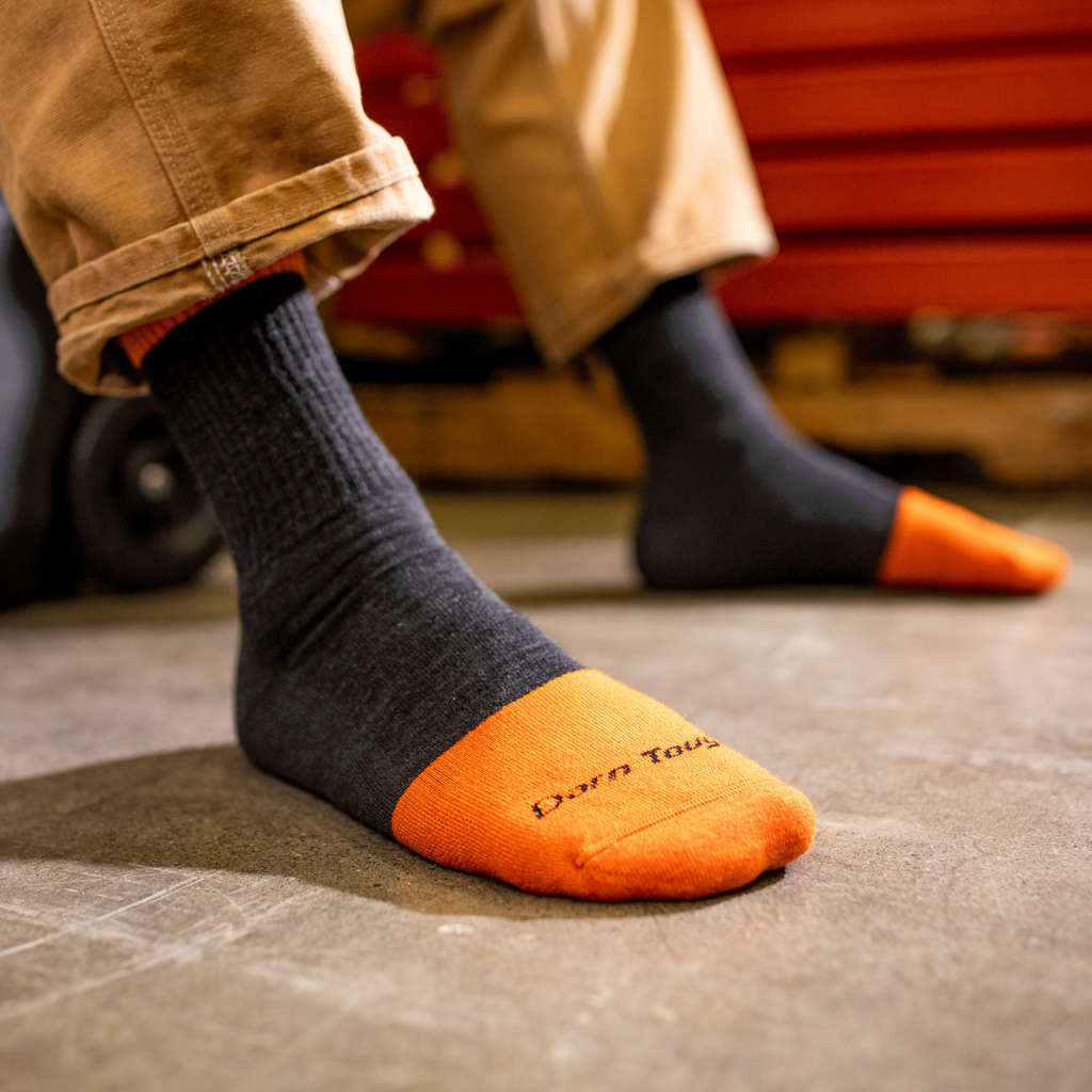 Person wearing dark graphite crew socks with orange reinforced toe cap in workshop setting