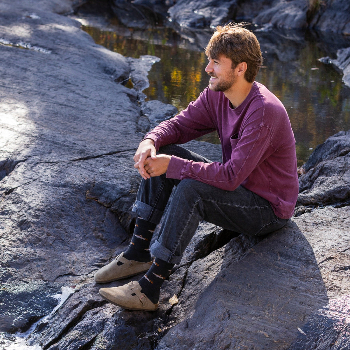 Man sitting on rocks near water wearing dark patterned crew socks with suede clogs