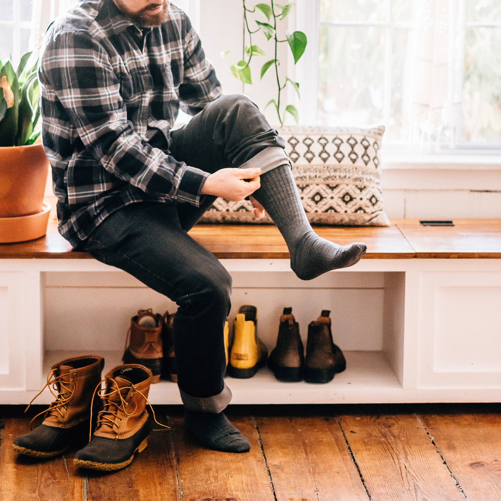 Man sitting on entryway bench pulling on gray mid-calf dress socks over dark pants