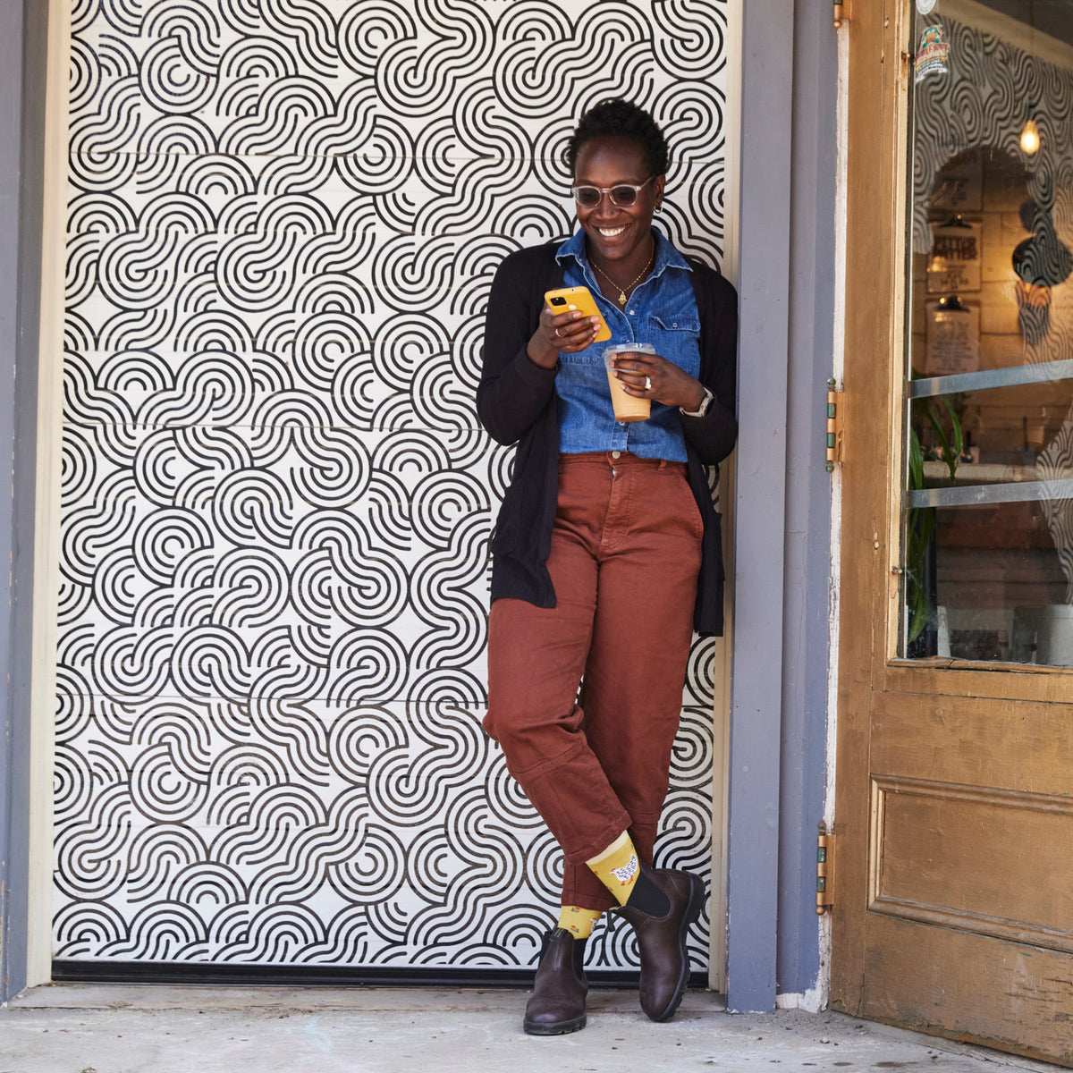 Woman leaning against patterned wall wearing yellow Darn Tough crew socks with ankle boots
