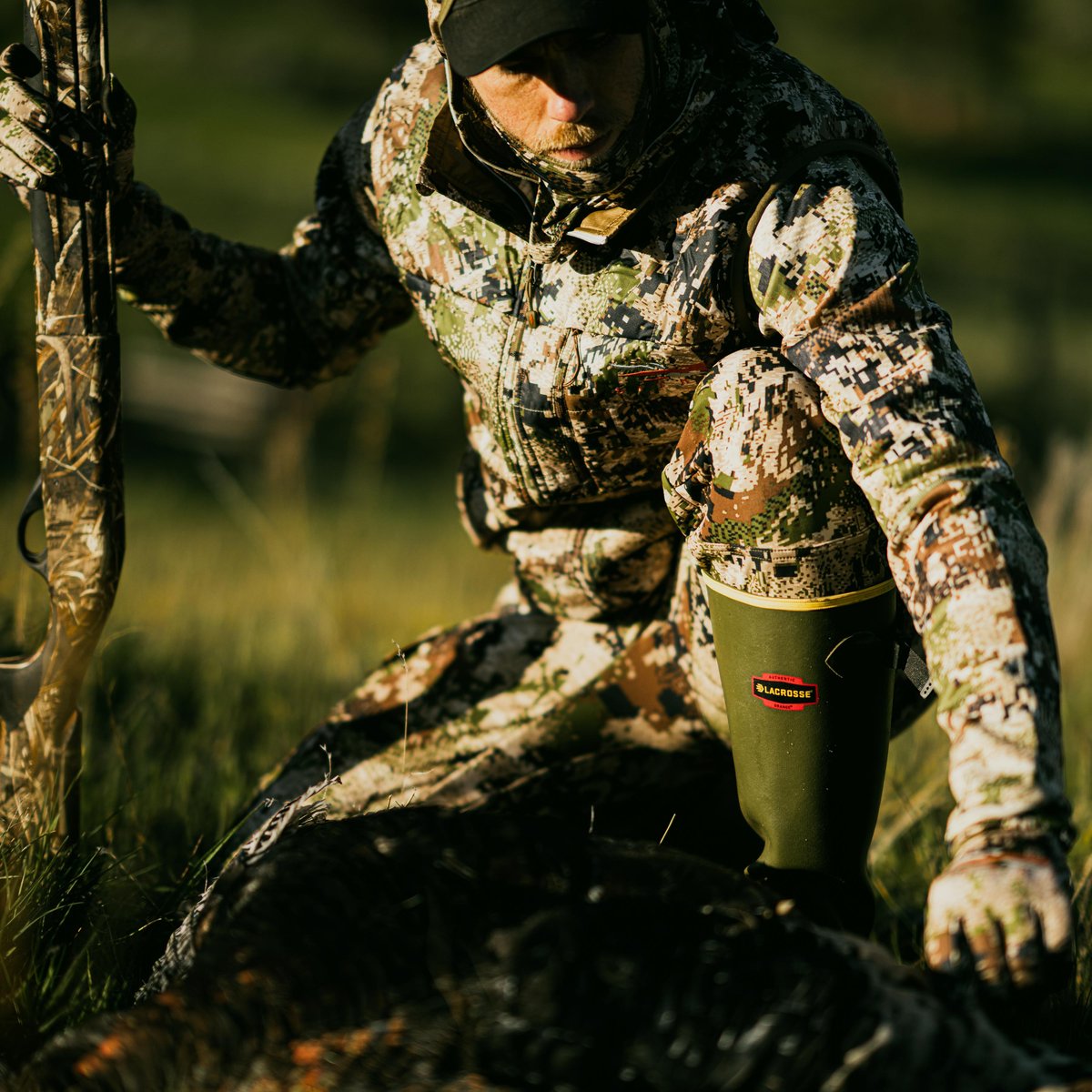 Hunter kneeling outdoors wearing camouflage gear and LaCrosse tall rubber boots