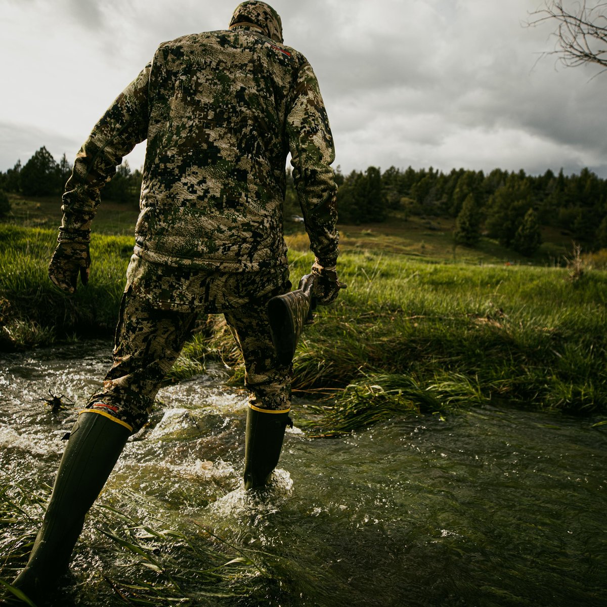 Person in camouflage wading through a stream wearing tall dark green rubber boots