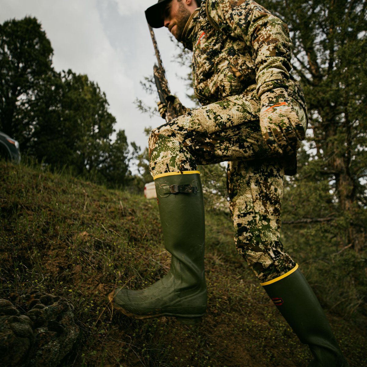 Hunter wearing tall olive green rubber boots with yellow trim on a wooded hillside