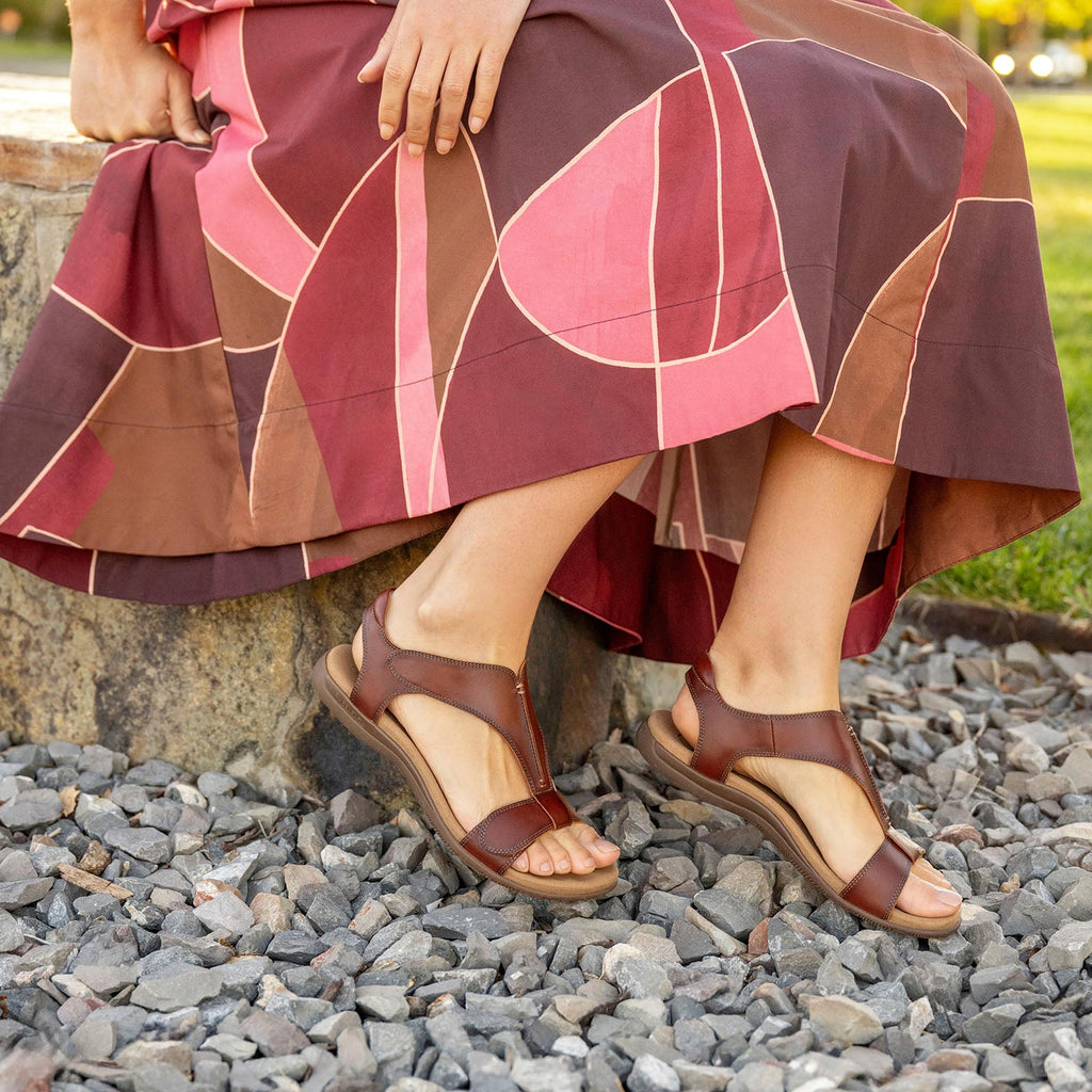 Woman seated outdoors wearing brown leather T-strap sandals on rocky ground with patterned skirt